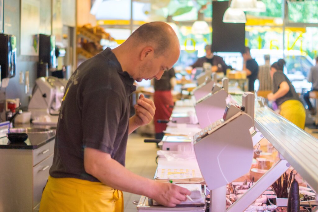 Ein Mann hinter einer Theke in einem Geschäft trägt eine gelbe Schürze und schreibt etwas auf, während weitere Personen unscharf im Hintergrund arbeiten.