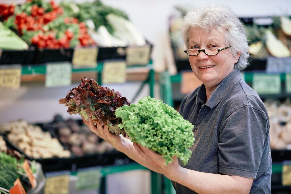 Ältere Frau mit Brille hält zwei verschiedene Salatköpfe in einem Gemüseladen und lächelt in die Kamera.