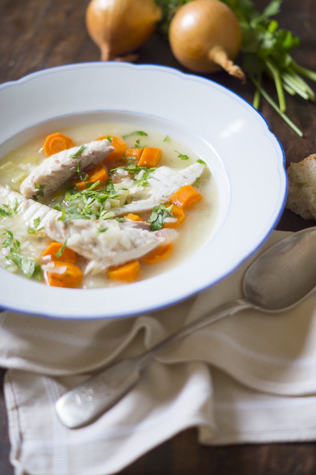Ein Teller Hühnersuppe mit Karotten, Petersilie und Hühnchenstücken, daneben Zwiebeln, frische Kräuter und ein Stück Brot auf einem Holztisch.