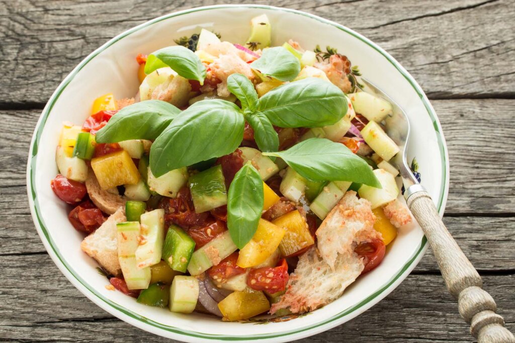 Bunter Brotsalat mit Tomaten, Gurken, Paprika, Zwiebeln, Brotstücken und frischem Basilikum in einer Schale, auf einem Holztisch serviert.