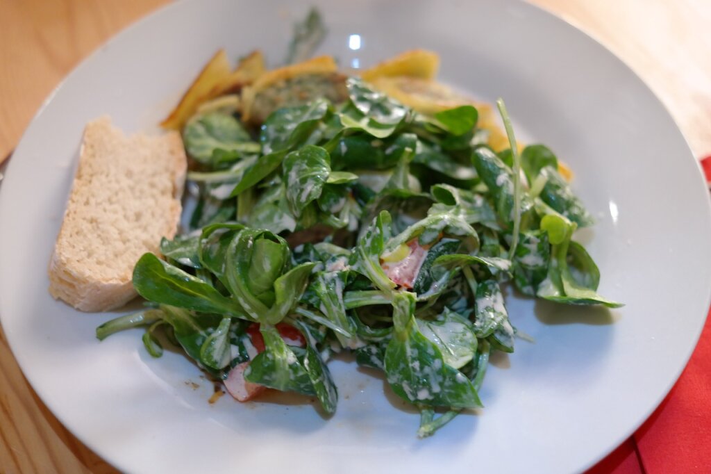 Ein Teller mit Feldsalat in Dressing, einem Stück Brot und gebratenen Kartoffeln.