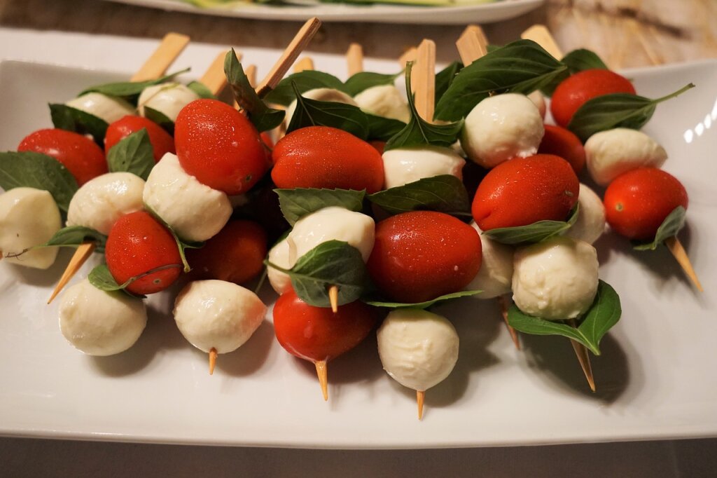 Mehrere Holzspieße mit kleinen Tomaten, Mozzarellakugeln und frischen Basilikumblättern auf einem weißen Teller.