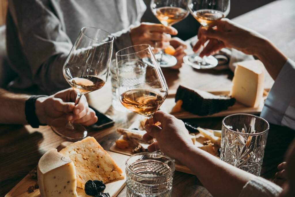 Eine Weinprobe mit Gläsern, die angestoßen werden. Ein Tisch mit verschiedenen Käsen im Hintergrund.