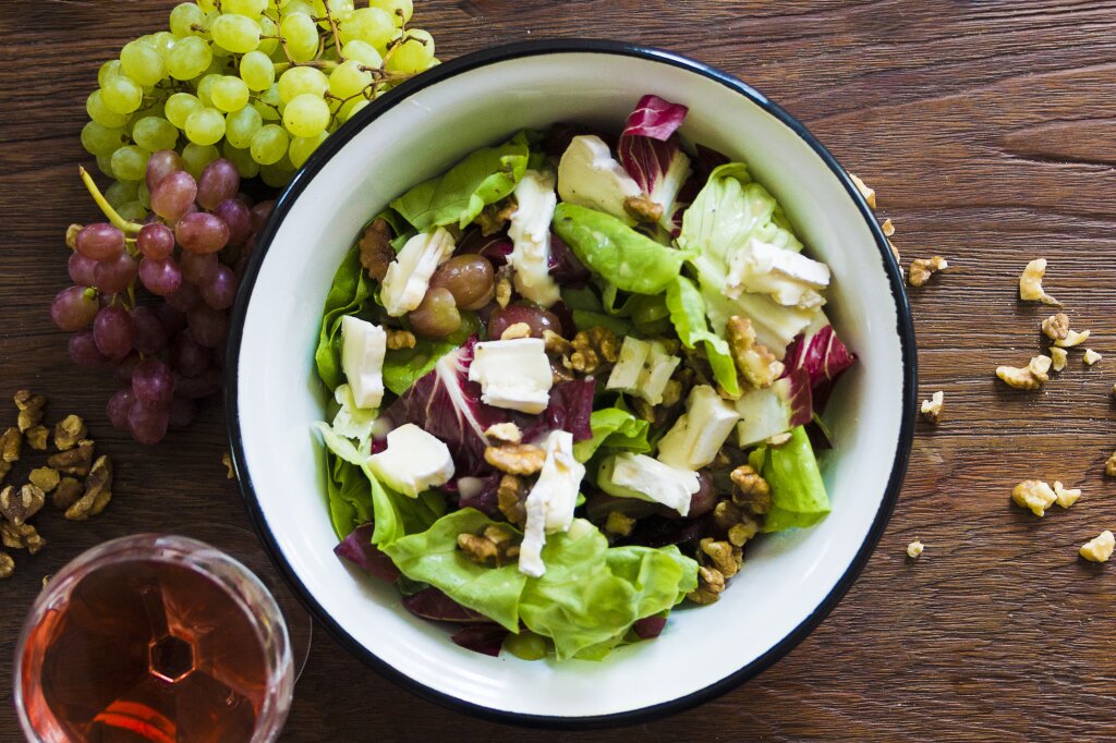 Ein Teller mit gemischtem Blattsalat, Trauben, Walnüssen und Weißschimmelkäse auf einem Holztisch, daneben ein Glas Roséwein und Trauben.