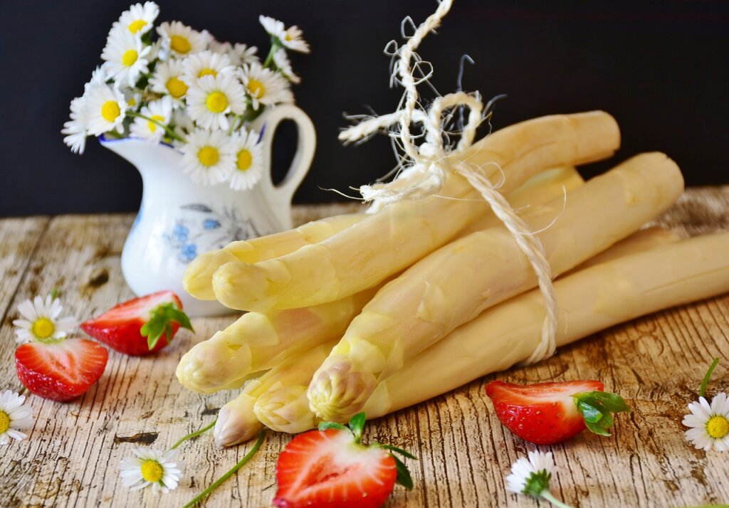 Ein Bund weißer Spargel, umgeben von aufgeschnittenen Erdbeeren und Gänseblümchen, im Hintergrund eine kleine Vase mit Gänseblümchen auf einem Holztisch.