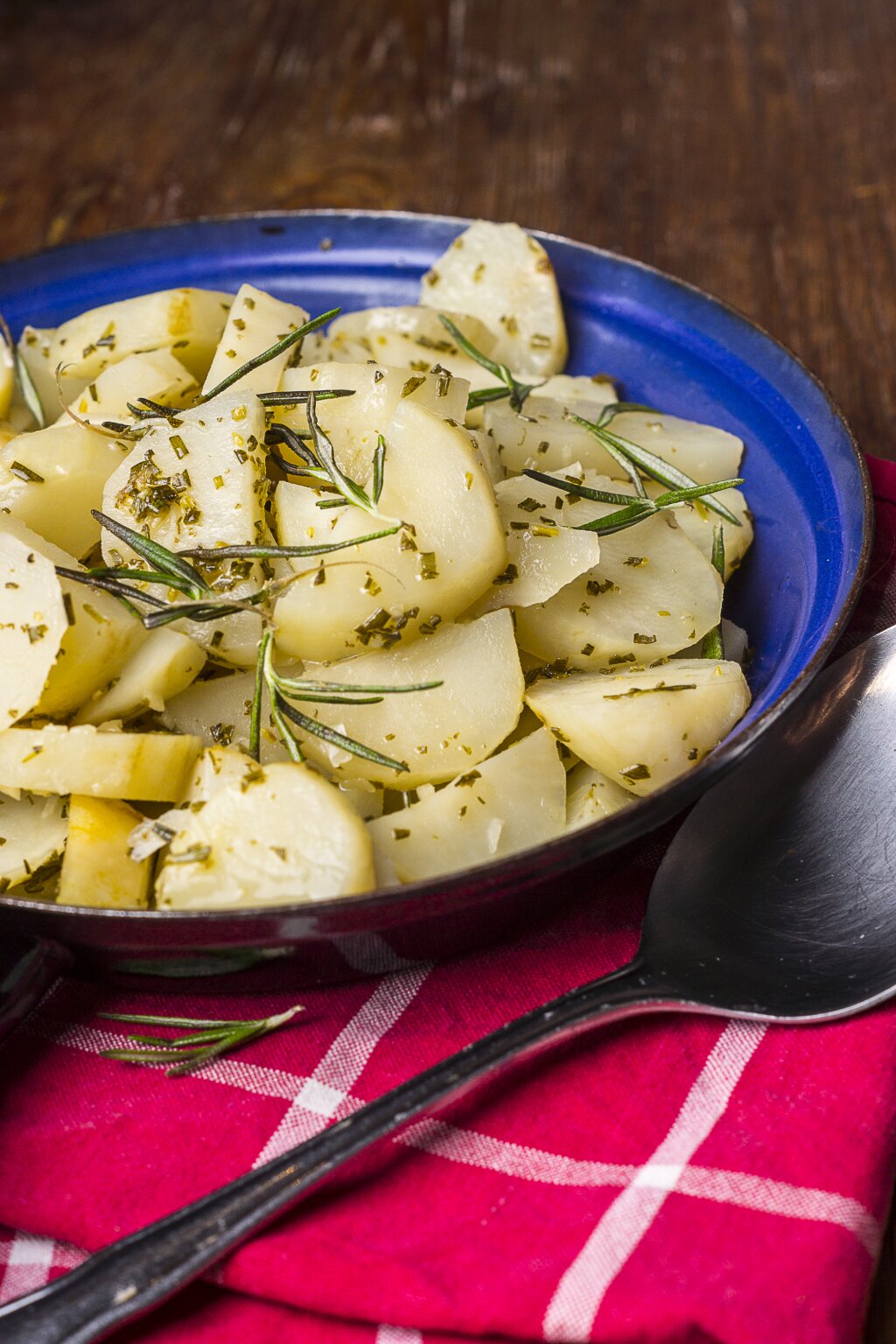 Gekochte Kartoffelscheiben mit frischen Kräutern, serviert in einer blauen Schüssel auf einem roten, karierten Tuch.