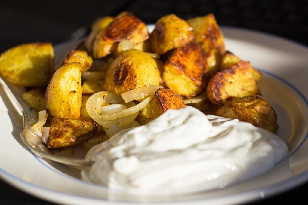 Gebratene Kartoffelstücke mit Zwiebeln auf einem Teller, daneben ein Klecks weißer Quark- oder Joghurtsauce.