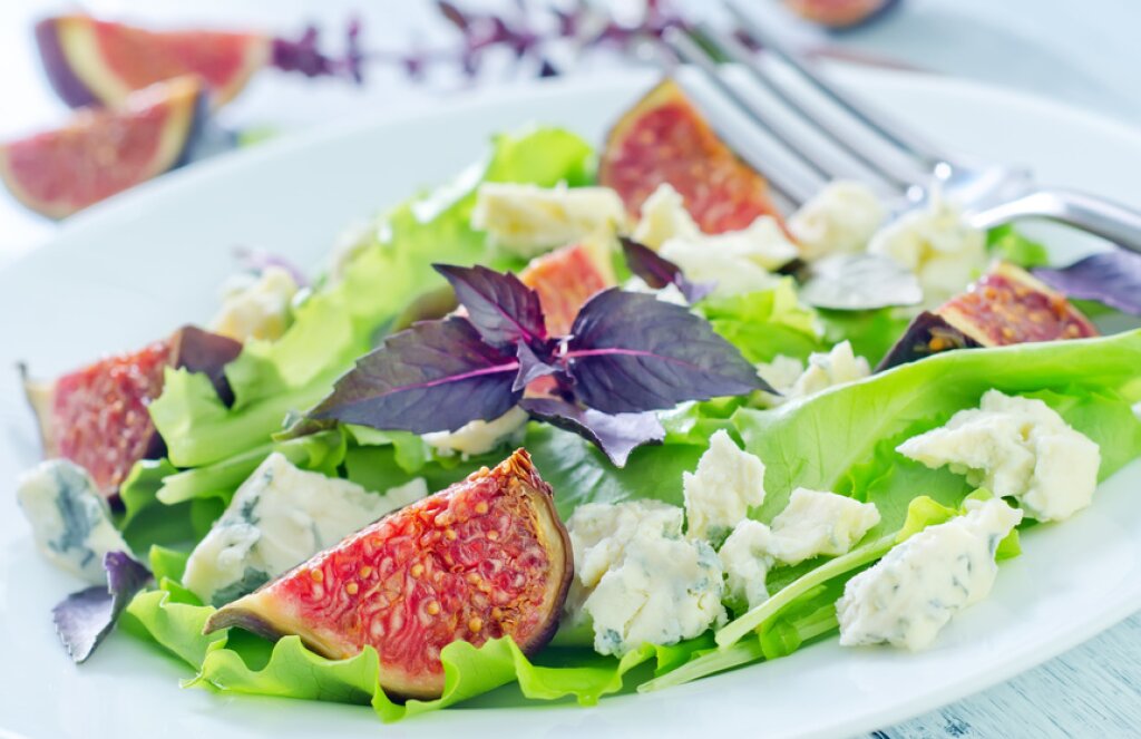 Salat aus grünen Blättern mit Feigen, Blauschimmelkäse und lila Basilikumblättern auf einem weißen Teller, im Hintergrund eine Gabel.