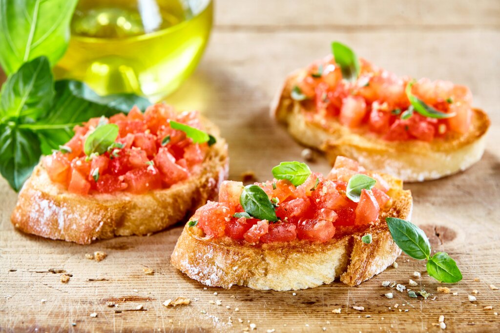 Drei Scheiben geröstetes Brot mit gewürfelten Tomaten, frischem Basilikum und Kräutern auf einem Holzbrett, im Hintergrund eine Flasche Olivenöl und Basilikumblätter.
