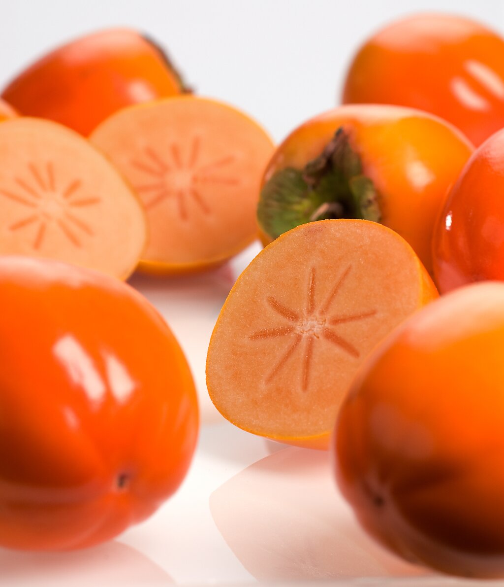 Mehrere ganze und aufgeschnittene reife Kakifrüchte mit orangefarbener Schale und glattem Fruchtfleisch, auf weißem Hintergrund.