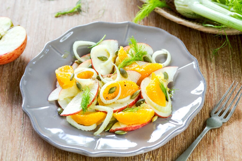 Ein Teller mit frischem Salat aus Apfelscheiben, Orangenspalten, Fenchel, Kürbiskernen und Fenchelgrün auf einem Holztisch, daneben eine Gabel.