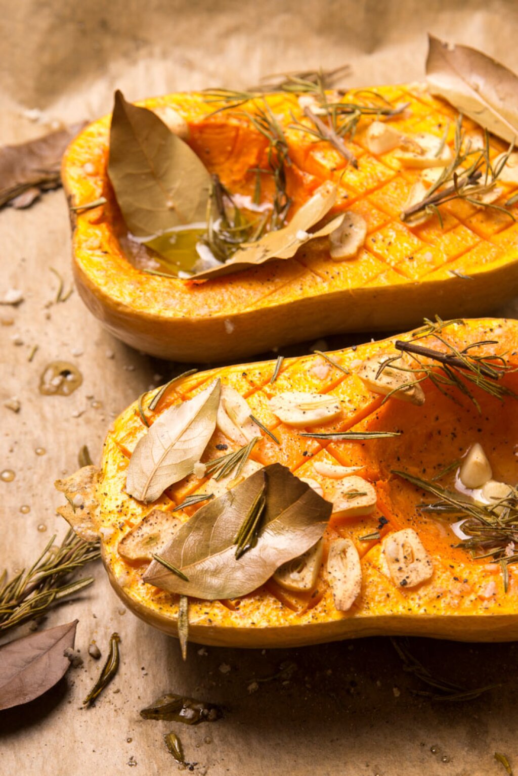 Zwei Hälften eines Butternut-Kürbisses, eingeritzt und belegt mit Knoblauchscheiben, Rosmarin, Lorbeerblättern, Salz, Pfeffer und Olivenöl auf Backpapier.