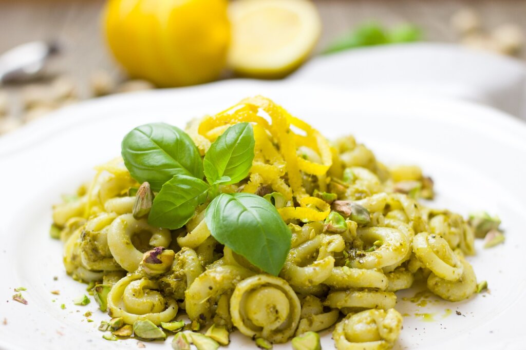Ein Teller Pasta mit grünem Pesto, Pistazien, Zitronenzesten und frischen Basilikumblättern, im Hintergrund eine angeschnittene Zitrone.