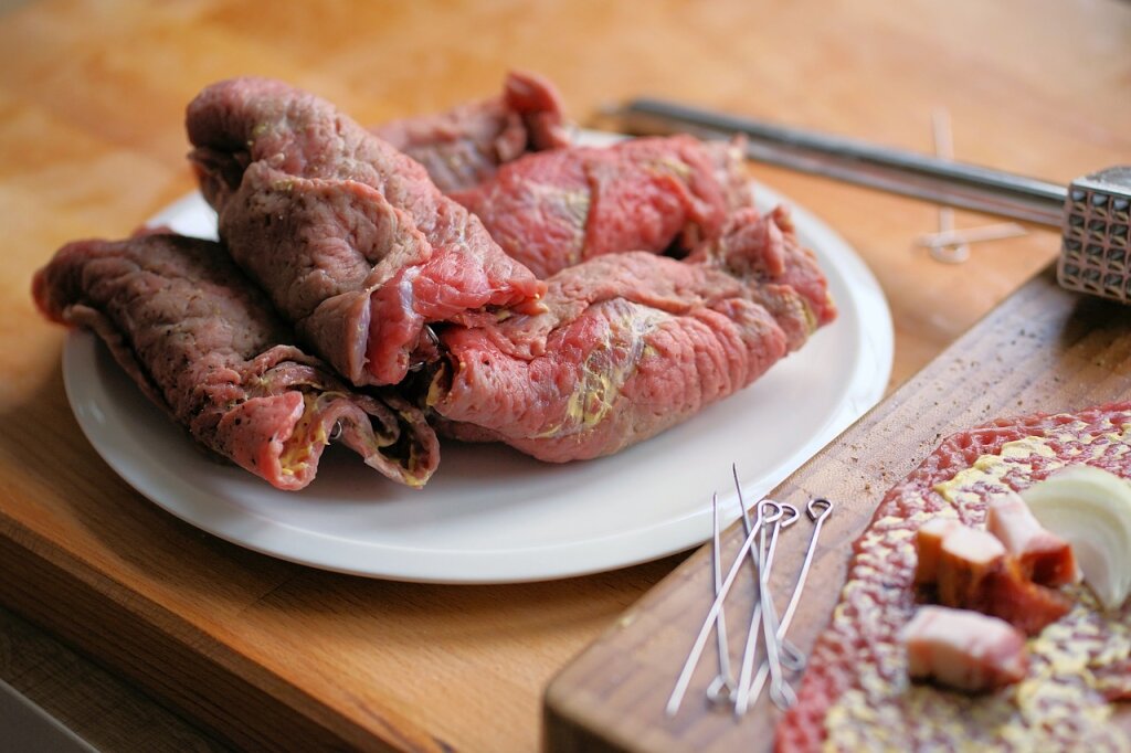 Mehrere ungekochte Rinderrouladen mit Füllung liegen auf einem weißen Teller, daneben Holzbrett mit Fleisch, Zwiebeln, Speck und Metallspieße.